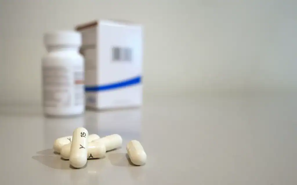 Close-up image of white capsules with medicine bottle and box in the background.