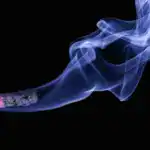 Detailed close-up of a cigarette with glowing ember and swirling smoke on a black background.