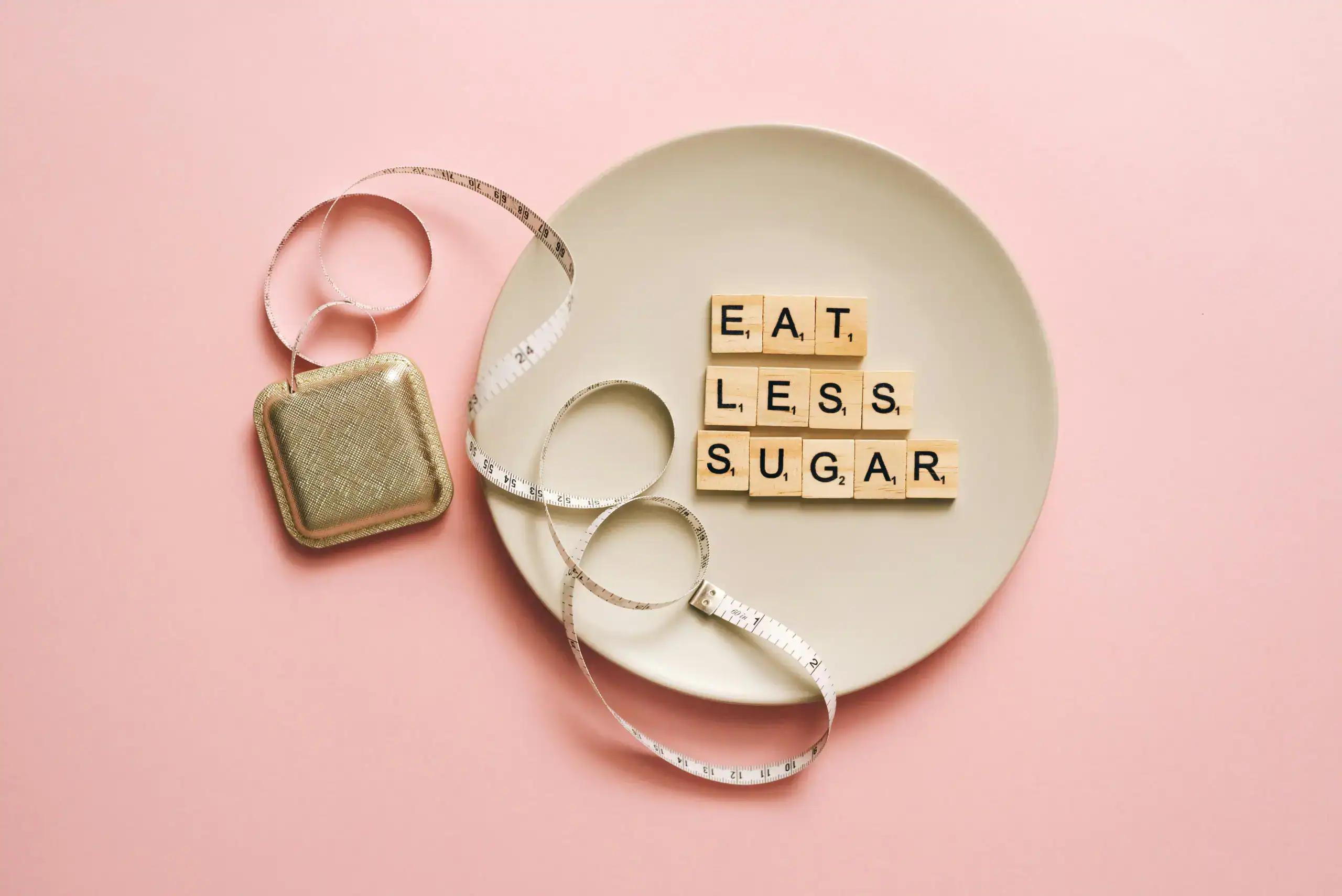 Flat lay of Scrabble tiles saying 'Eat Less Sugar' on a plate with a tape measure on a pink background.