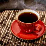 A steaming cup of coffee in a red mug surrounded by coffee beans on a bamboo mat.