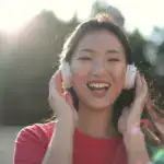 Smiling woman in red t-shirt listening to music with headphones in a sunny outdoor setting.
